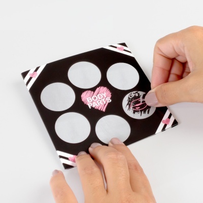 Paleta de maquilhagem preta com seis espaços vazios e corações rosa, com texto BODY PARTS e LIPS, segurada por mãos femininas