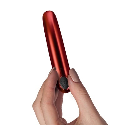 Vibrador vermelho metálico com botão preto em forma de coração segurado por mão feminina em fundo branco.