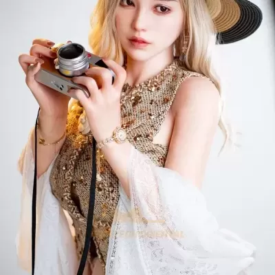 Mulher loira com chapéu de palha segurando uma câmera prateada vestida com blusa castanha de paetês e xale branco de renda