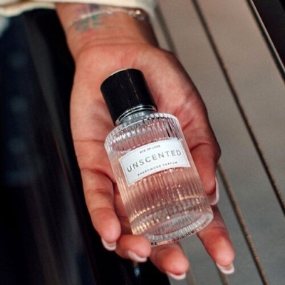 Frasco de perfume transparente com tampa preta e etiqueta branca com texto, segurado por uma mão com tatuagem