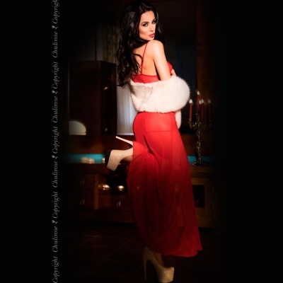 Modelo com vestido vermelho transparente, estola branca de pelo e sapatos dourados de salto alto num ambiente interior escuro.