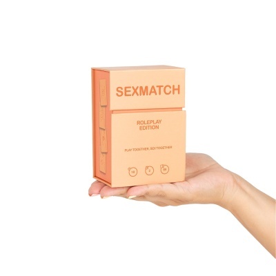 Caixa cor salmão com texto SEXMATCH e detalhes sobre o jogo na mão de uma pessoa.