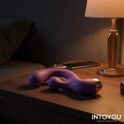Vibrador roxo curvo com base prateada numa mesa de madeira junto a telemóvel e lâmpada