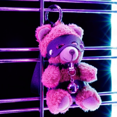 Urso de peluche cor de rosa com acessórios pretos pendurado em estrutura metálica.