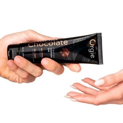Tubo preto de gel íntimo sabor chocolate com texto em dourado e branco, segurado por duas mãos.