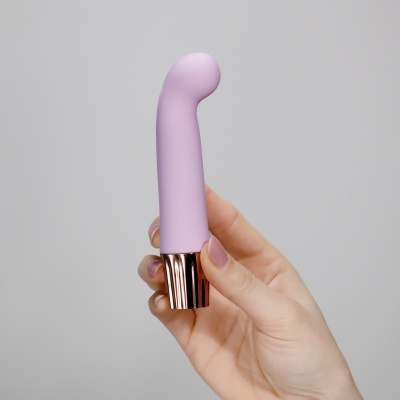 Vibromassageador lilás com base dourada numa mão com unhas cor de rosa.