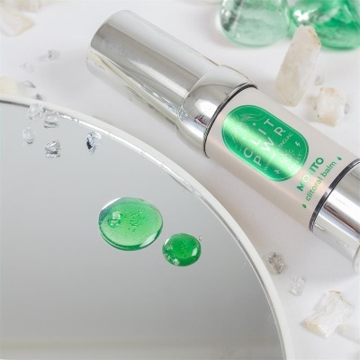 Frasco branco e prateado de creme com rótulo verde junto a pedras e gotas verdes num espelho