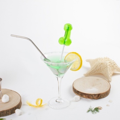 Cocktail verde em copo com rodela de limão, pirulito em forma de falo, canudo metálico, pedaços de madeira, pedras brancas e estrela-do-mar