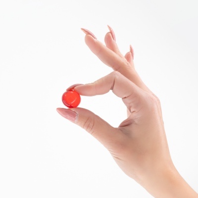 Mão segurando cápsula vermelha com unhas pintadas de rosa claro
