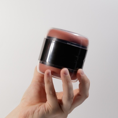 Pote de plástico com tampa transparente, corpo preto e produto castanho avermelhado, segurado por mão