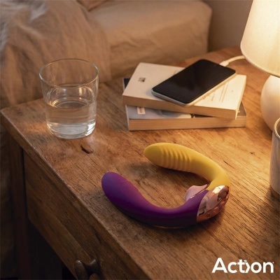 Aparelho vibrador em silicone roxo e amarelo com detalhes dourados numa mesa de madeira com copo de água, livros e telemóvel