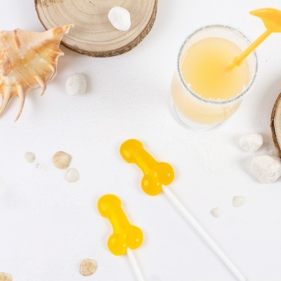 Dois rebuçados amarelos em forma de pênis com palitos junto a conchas, pedras e um copo com bebida amarela e borda salgada.