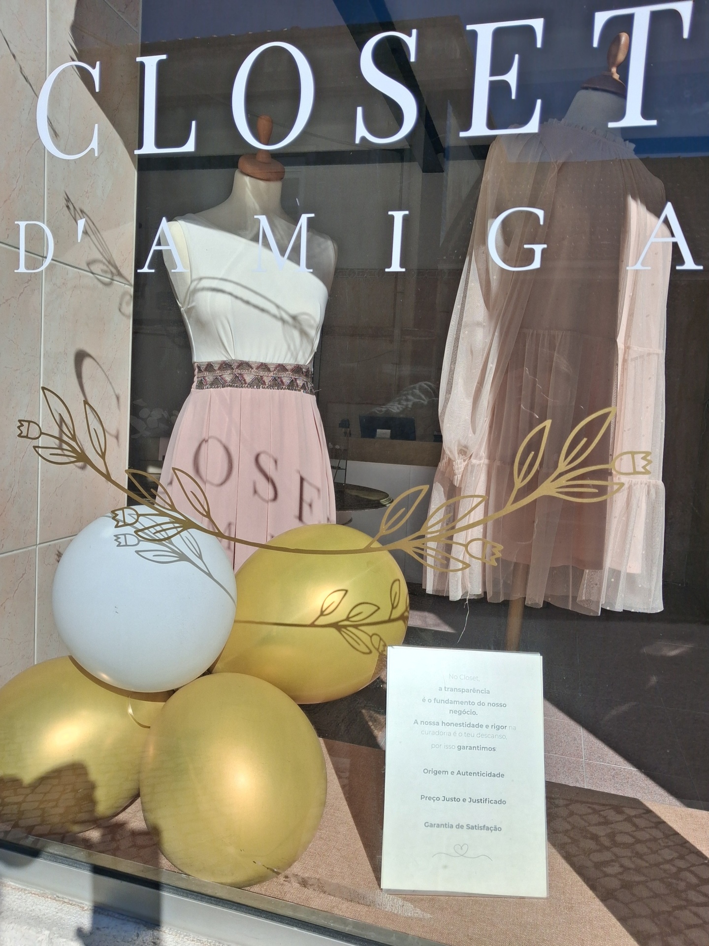 Vitrine de loja com vestidos pasteis em manequins e balões dourados e brancos
