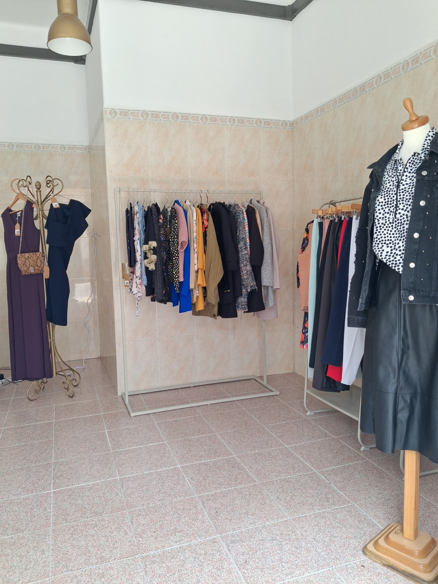 Roupas femininas diversas penduradas em cabides e manequim com camisa de bolinhas e casaco preto numa loja com paredes de azulejo e chão de mosaico.