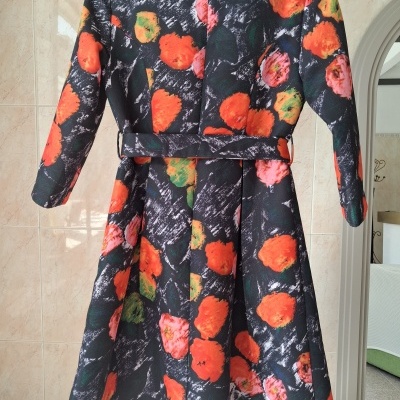 Casaco floral comprido preto com flores laranja e vermelho pendurado numa parede interior.