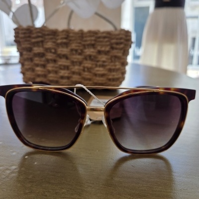 Óculos de sol com armação castanha e lente escura sobre mesa com bolsa de palha ao fundo