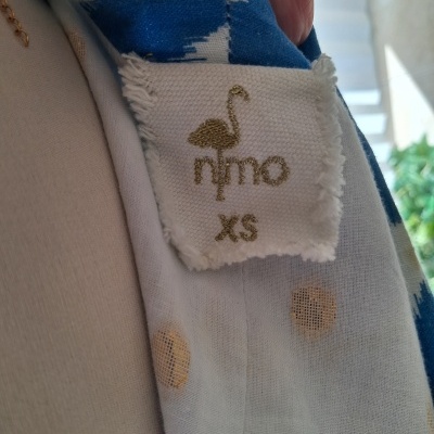 Etiqueta branca bordada a dourado com flamingo e texto nimo XS numa peça de tecido azul e branca com padrão e bolinhas.