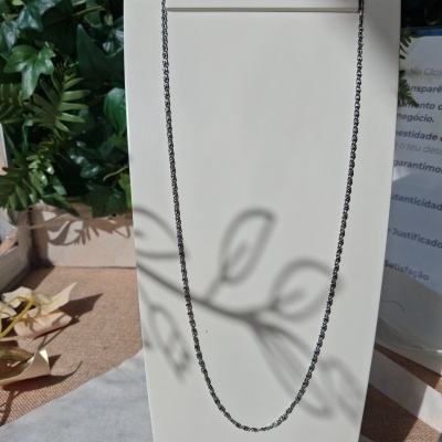 Colar fino de corrente metálica prateada num suporte branco com fundo de plantas.