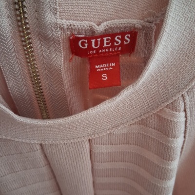 Etiqueta vermelha GUESS em peça de roupa cor de rosa clara com fecho dourado.