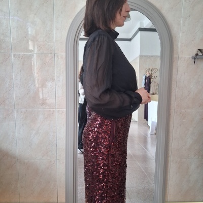 Mulher com camisa preta transparente e saia com lantejoulas vermelhas em frente a espelho com moldura cinza.