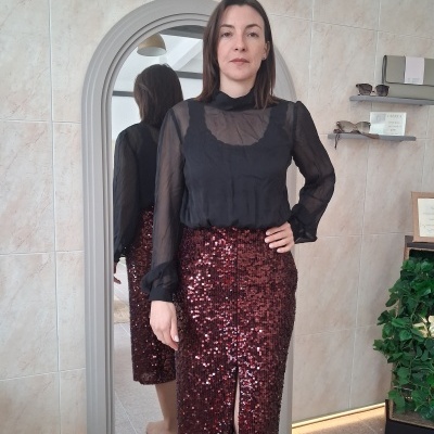 Mulher com blusa preta transparente e saia midi bordô com paetês vermelhos em frente a espelho