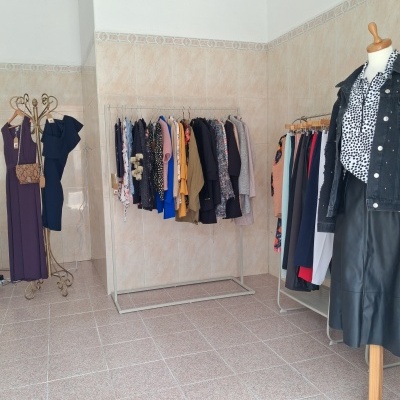 Roupas femininas diversas penduradas em cabides e manequim com camisa de bolinhas e casaco preto numa loja com paredes de azulejo e chão de mosaico.