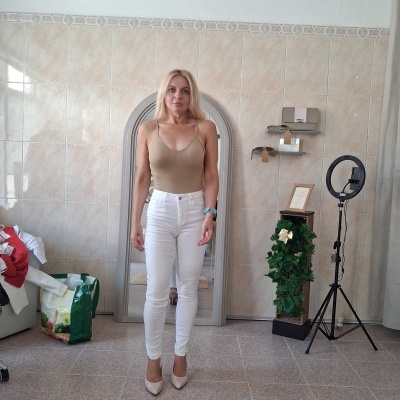 Mulher com top bege e calças brancas posando em frente a espelho e parede de azulejos bege