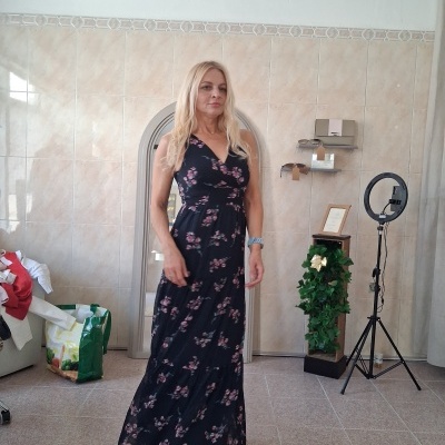Vestido longo preto com flores rosa e verde, usado por mulher em quarto com parede e chão de azulejos.