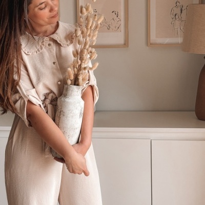 Mulher com macacão bege segurando vaso cerâmico branco com flores secas em sala com decoração neutra