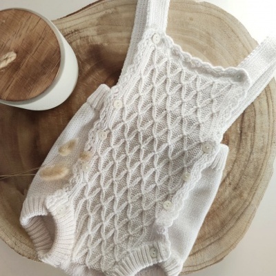 Jardineira de bebé branca em malha texturizada com botões, sobre base de madeira clara