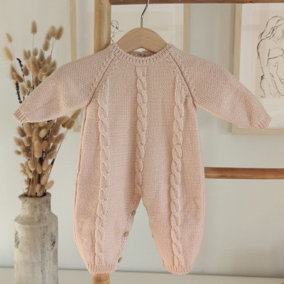 Macacão de bebé rosa claro em malha com padrão de tranças pendurado num cabide de madeira