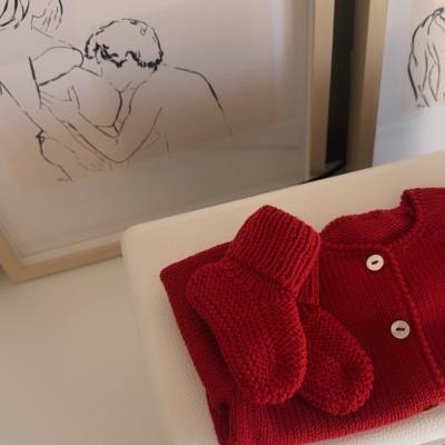 Roupa de bebé vermelha de malha com botões sobre móvel branco com quadros ilustrados ao fundo.