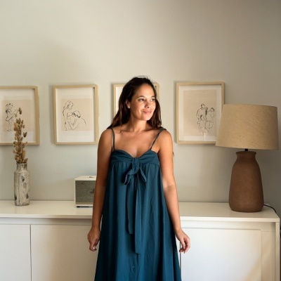 Mulher veste vestido azul comprido de alças finas em interior moderno com móveis brancos e decoração minimalista.