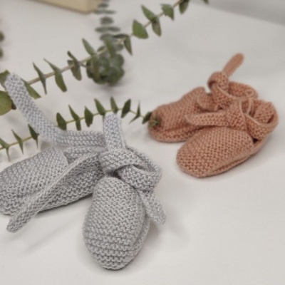 Dois pares de sapatinhos de bebé tricotados, cinza e rosa, com laços, sobre superfície branca com folhagem ao fundo.