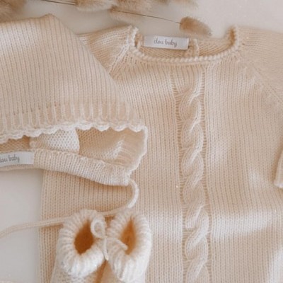Conjunto de roupa de bebé creme de malha com body, gorro e sapatinhos ao lado de ramagem secada