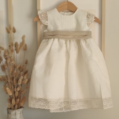 Vestido infantil branco com renda e fita bege pendurado em cabide de madeira