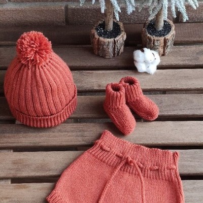 Conjunto de bebé cor de terracota com gorro, botinhas e calções em malha, em madeira com árvores decorativas ao fundo