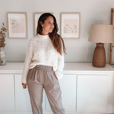 Mulher vestindo suéter branco de malha e calças bege em sala decorada com móvel branco, quadros e candeeiro castanho