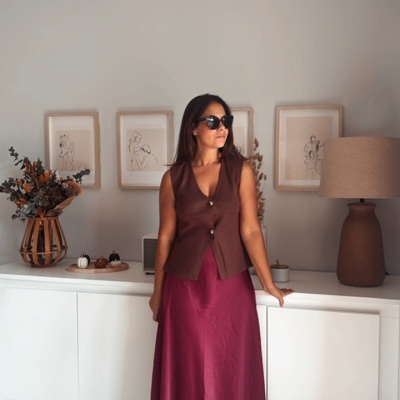 Mulher com vestido vinho e colete castanho junto a móvel branco com decoração minimalista.