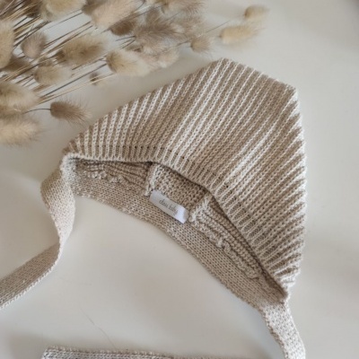 Gorro de malha listrado bege claro e branco com tiras para amarrar e plantas secas ao fundo.