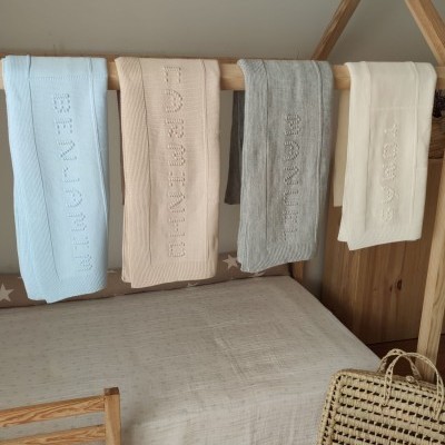 Quatro mantas de bebé coloridas penduradas numa cama infantil de madeira clara com uma cesta de palha ao lado