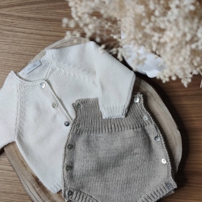 Conjunto de roupa de bebé em malha com camisola branca e calções cinzentos sobre base de madeira com flores secas
