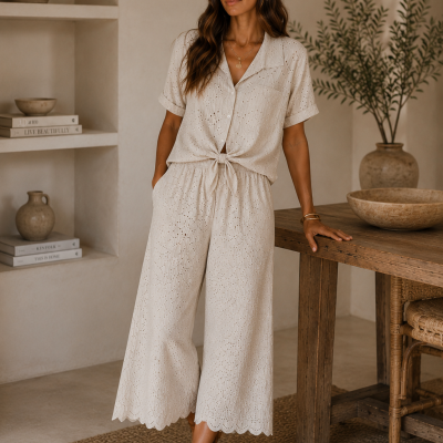 Conjunto feminino bege com camisa de manga curta com nó e calças largas bordadas, sandálias castanhas, cenário neutro com mesa de madeira e vasos de cerâmica.
