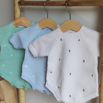 Bodies de bebé de malha verdes, azuis e rosa claro com pontos, pendurados em cabides de madeira.