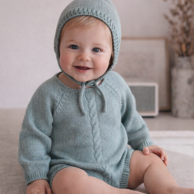 Bebé com camisola e gorro de malha azul clara sentado num sofá cinza claro.