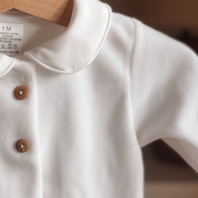 Body branco para bebé com gola redonda e botões de madeira pendurado em cabide de madeira