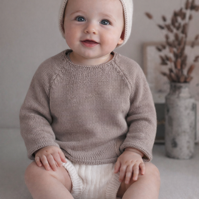 Bebé com roupa de malha em tons neutros sentado em superfície clara com vaso cinzento ao fundo