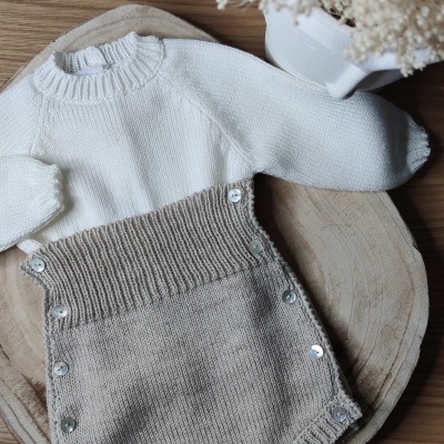 Conjunto de roupa de bebé em malha branca e bege com botões prateados, sobre base de madeira com vaso e flores secas