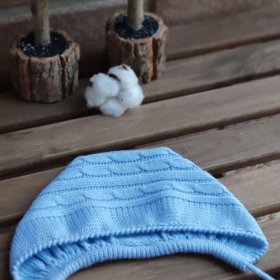 Gorro azul claro de malha com cordão sobre mesa de madeira, ao fundo duas árvores decorativas e ramo de algodão.