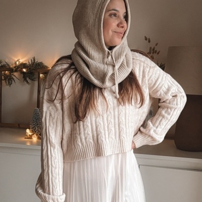 Mulher com capuz de malha bege e camisola creme numa sala decorada para Natal.